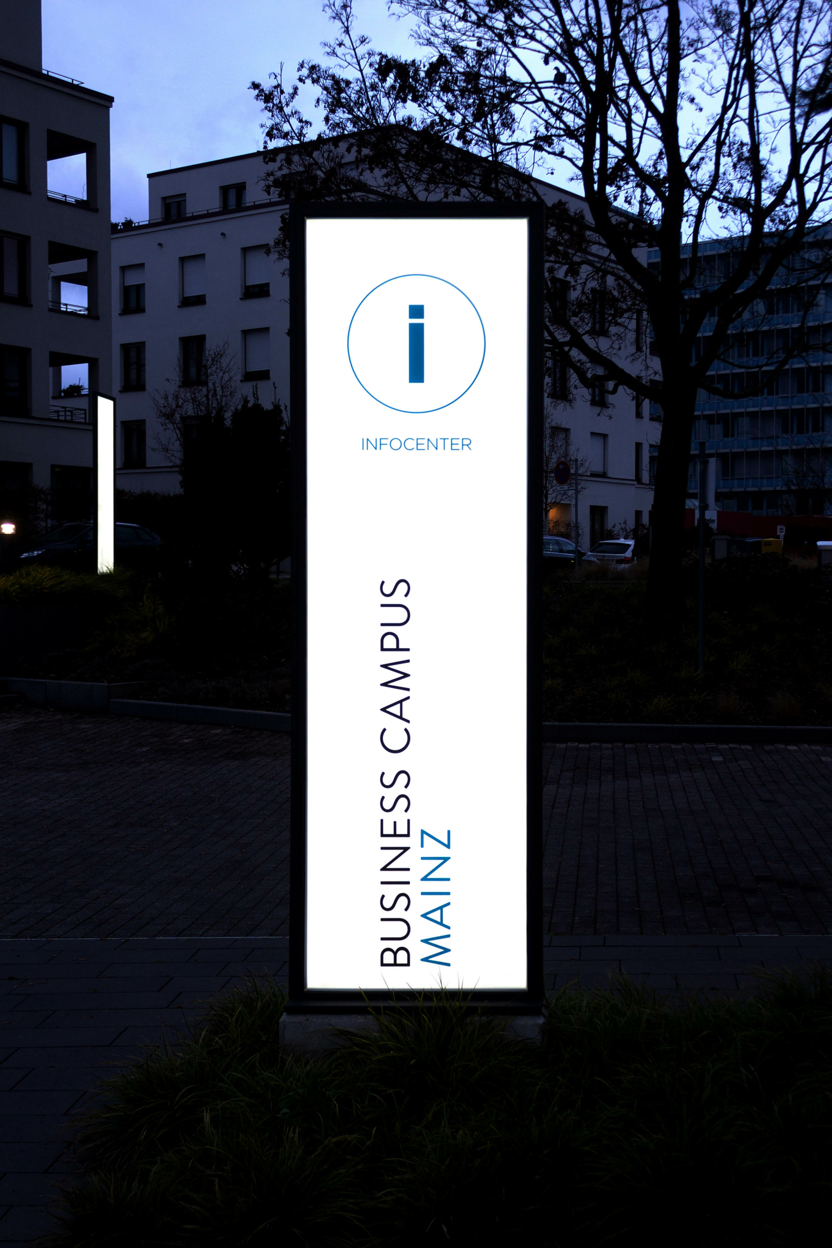 Werbepylon, beleuchtet, als LED-Firmenschild aus Glas auf dem Business Campus Mainz. Die weiße Leuchtstele erhellt gleichzeitig den Vorplatz.
