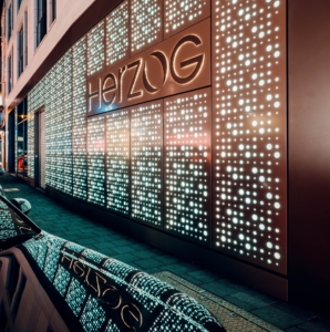 Outdoor LED-Panels hinter einer bronze-farbenen Lochblende mit runden Löchern und Schriftzug "Herzog" als Fassadenbeleuchtung in Köln.