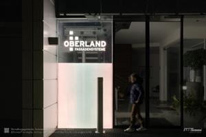 LED-Panel als Outdoor-Beleuchtung aus Glas eines Firmen-Eingangs: Unten matt leuchtend, oben klar mit leuchtendem Logo.